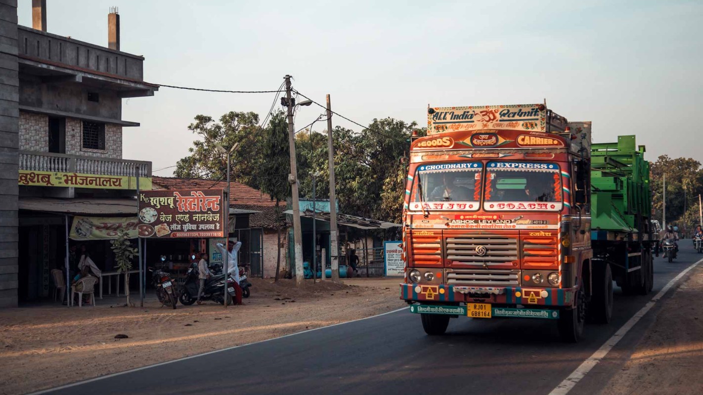 A vibrant truck travels along a road in the countryside of Maharashtra, with local shops lining the street and warm sunlight casting soft shadows in the evening.