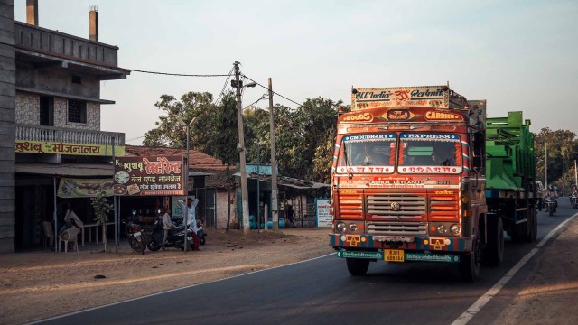 A vibrant truck travels along a road in the countryside of Maharashtra, with local shops lining the street and warm sunlight casting soft shadows in the evening.
