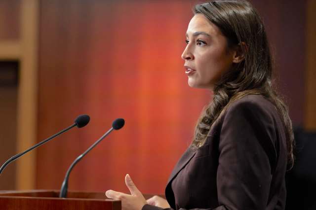 Rep. Alexandria Ocasio-Cortez, in a brown blazer, speaks at a podium with microphones against a blurred background.