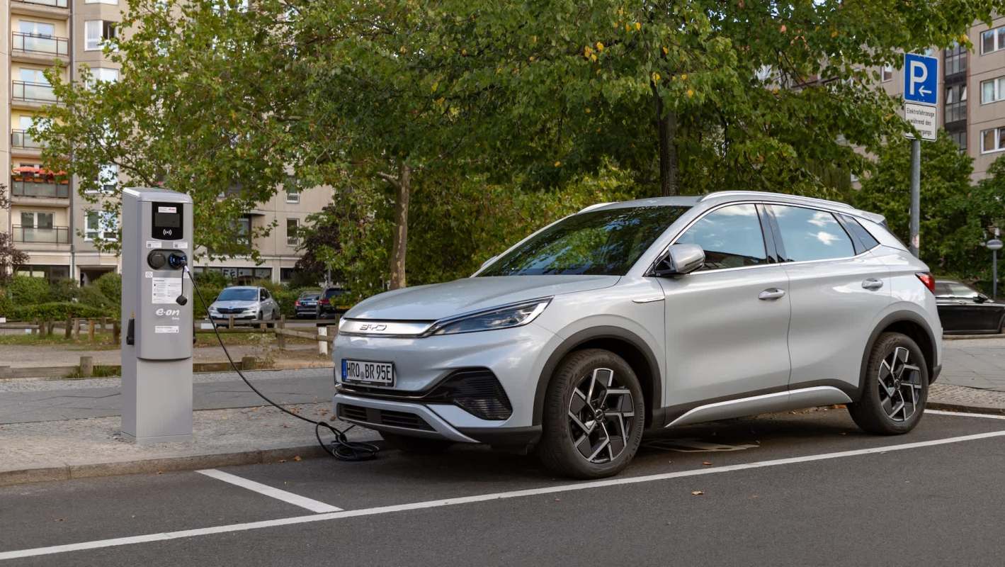 A silver electric vehicle plugged into a charging station on a city street with green trees in the background.