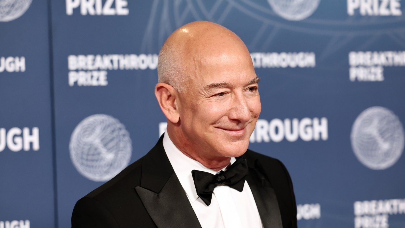 Jeff Bezos, in a tuxedo, smiles at the camera, standing in front of a blue backdrop with "Breakthrough Prize" logos.