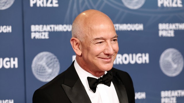 Jeff Bezos, in a tuxedo, smiles at the camera, standing in front of a blue backdrop with "Breakthrough Prize" logos.