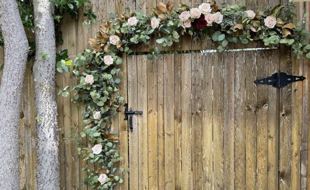 Two tree trunks next to a wooden garden gate adorned with flowers and greenery.