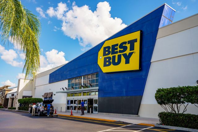 A Best Buy store with a blue exterior and a large yellow sign on a sunny day, featuring customers near a car.