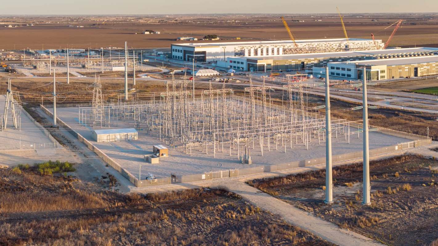 An aerial view of a power substation with transmission towers and a construction site in the background.