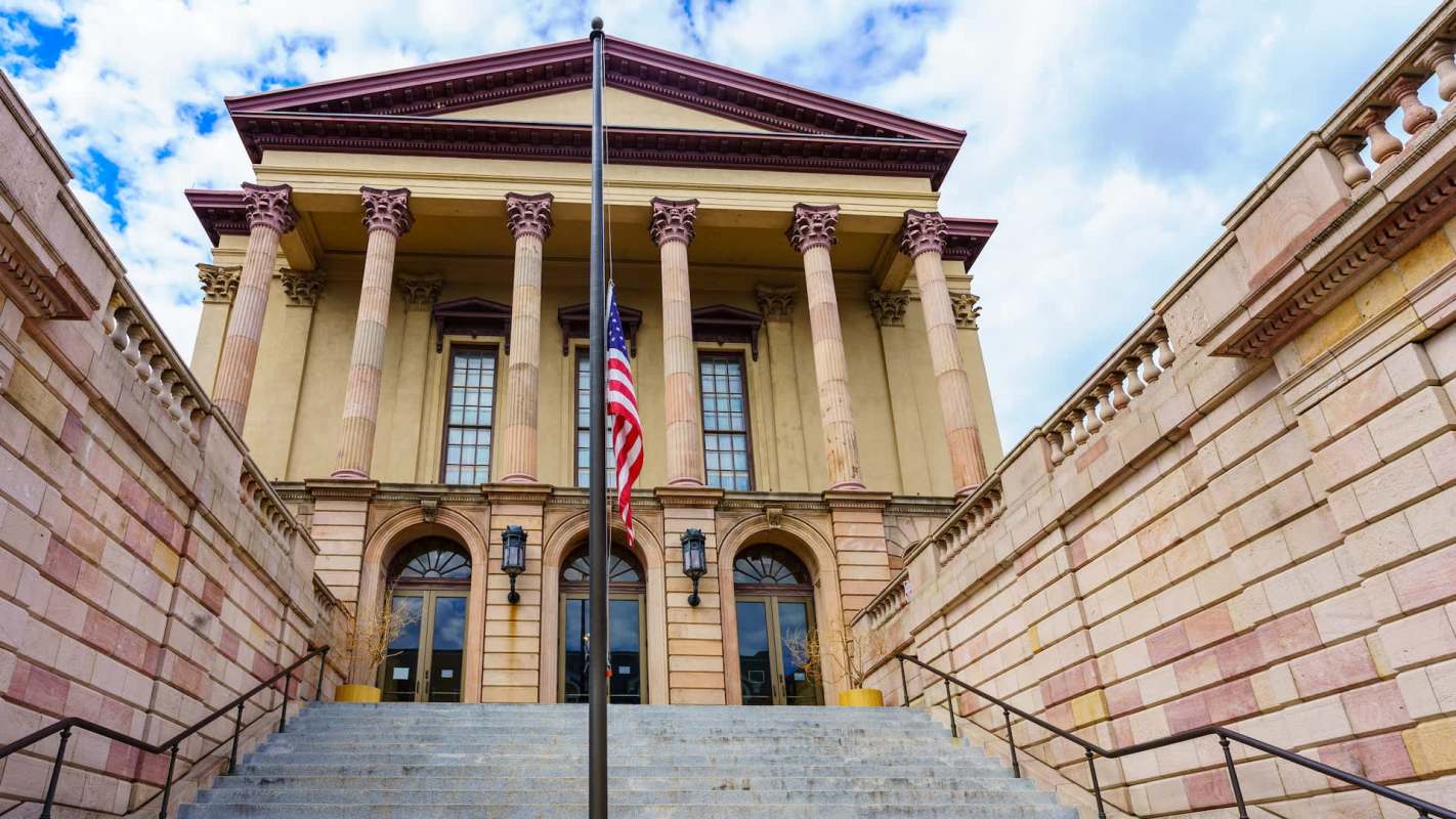 A grand building with columns, an American flag, and steps leading up to the entrance under a blue sky.