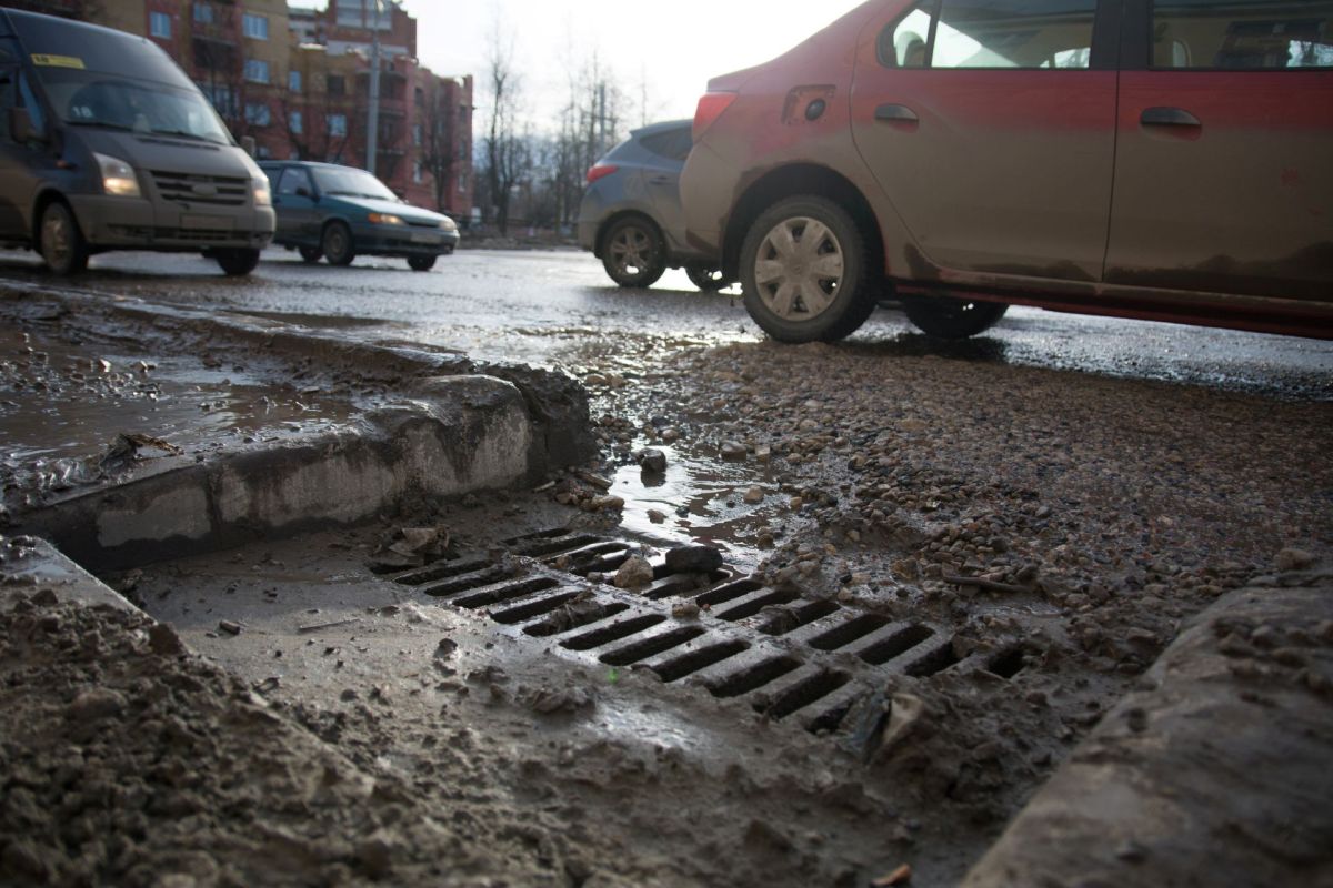 A muddy street with water pooling near a drainage grate, flanked by parked and passing vehicles.