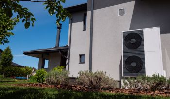 A modern house exterior featuring an outdoor heat pump unit amidst greenery and blue sky.