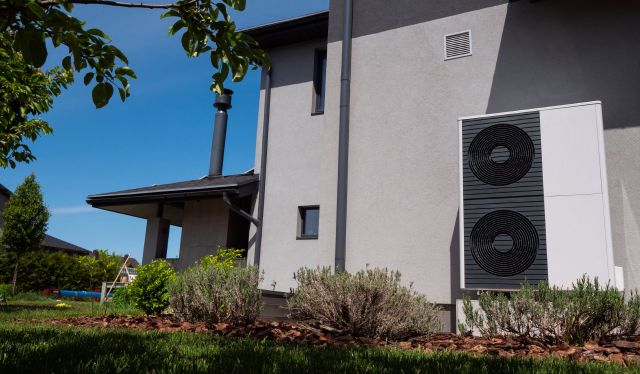A modern house exterior featuring an outdoor heat pump unit amidst greenery and blue sky.
