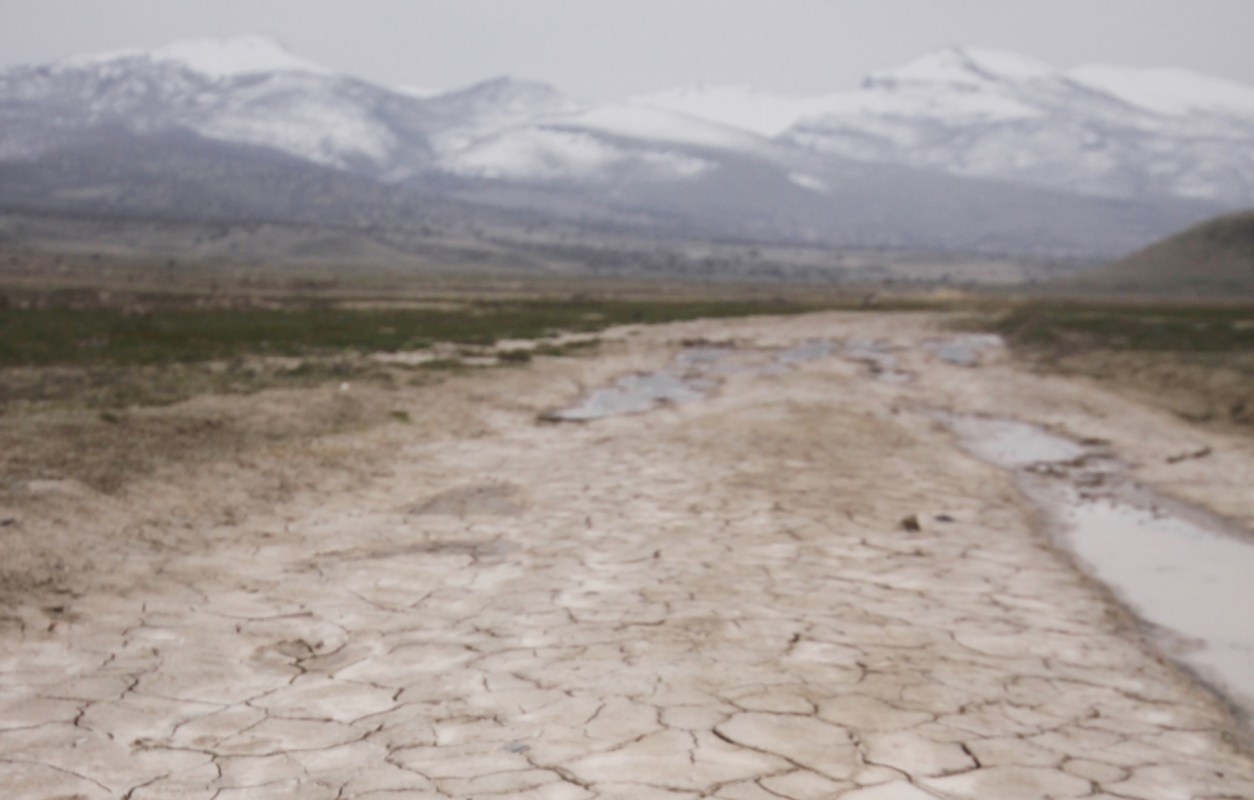 A dry, cracked landscape stretches toward snow-capped mountains under an overcast sky.