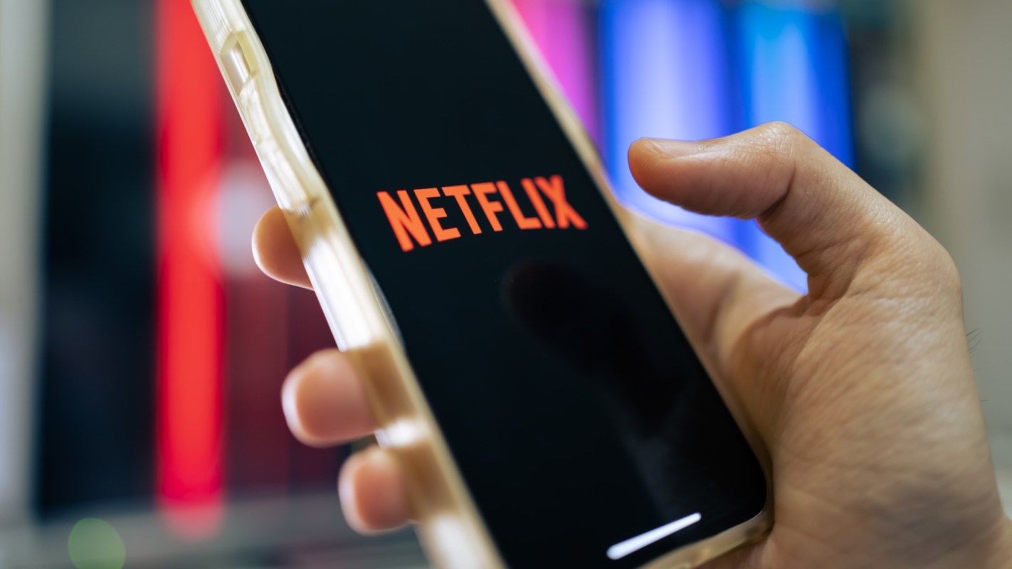 A hand holds a smartphone displaying the Netflix logo with colorful lights blurred in the background.