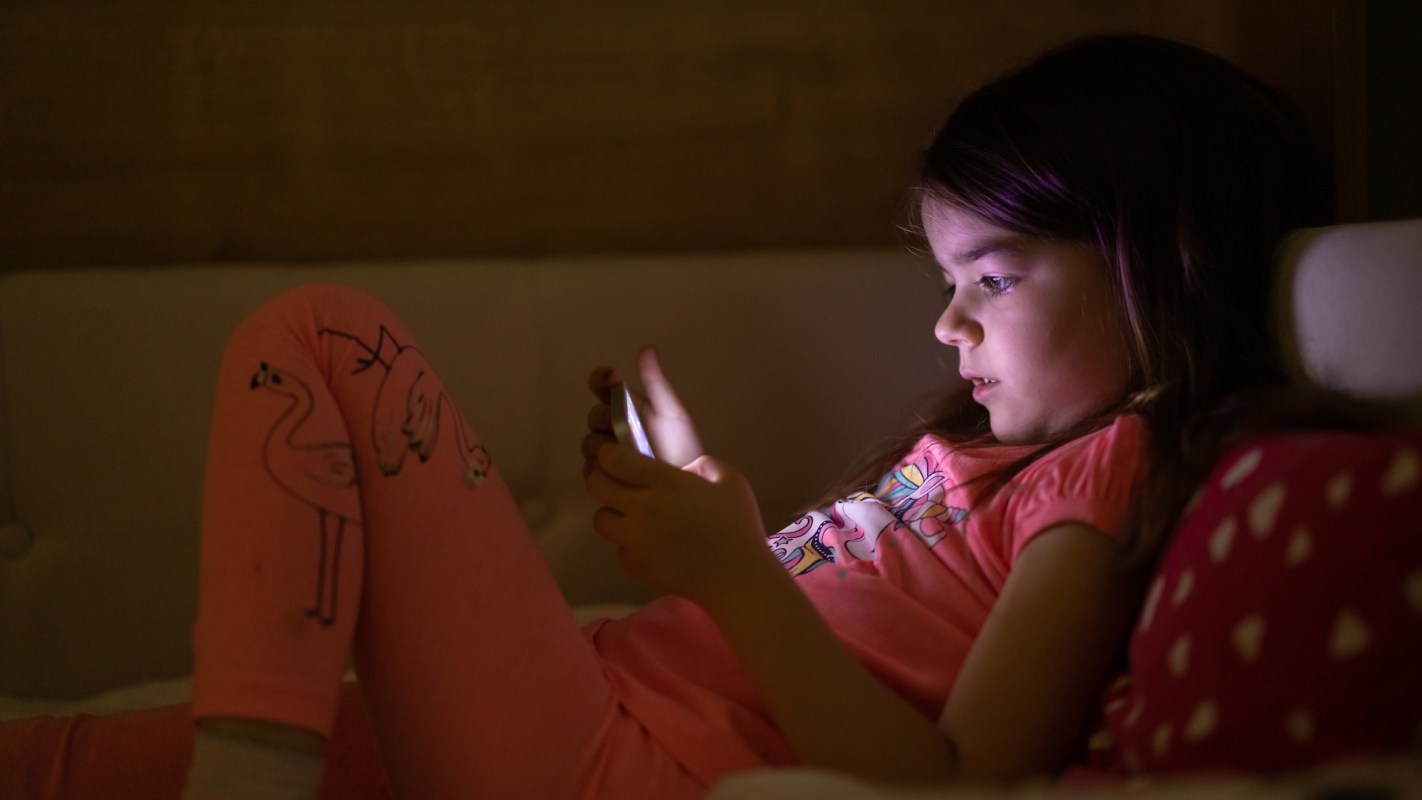 A girl in pink pajamas lies on a couch, focused on her phone in a dimly lit room.