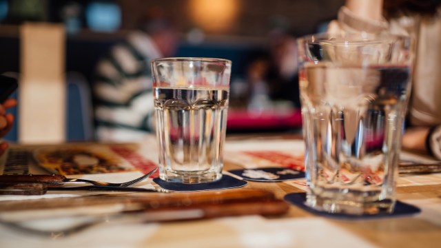 A close-up of a glass of water on a table with cutlery and blurred background elements.