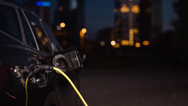 An electric vehicle at a charging station at night with city lights blurred in the background.