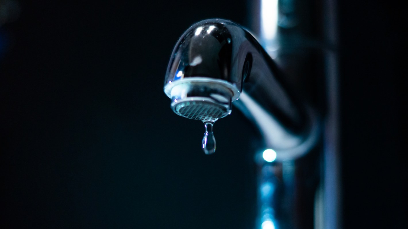 A close-up of a shiny faucet with a single droplet of water hanging from the tip against a dark background.