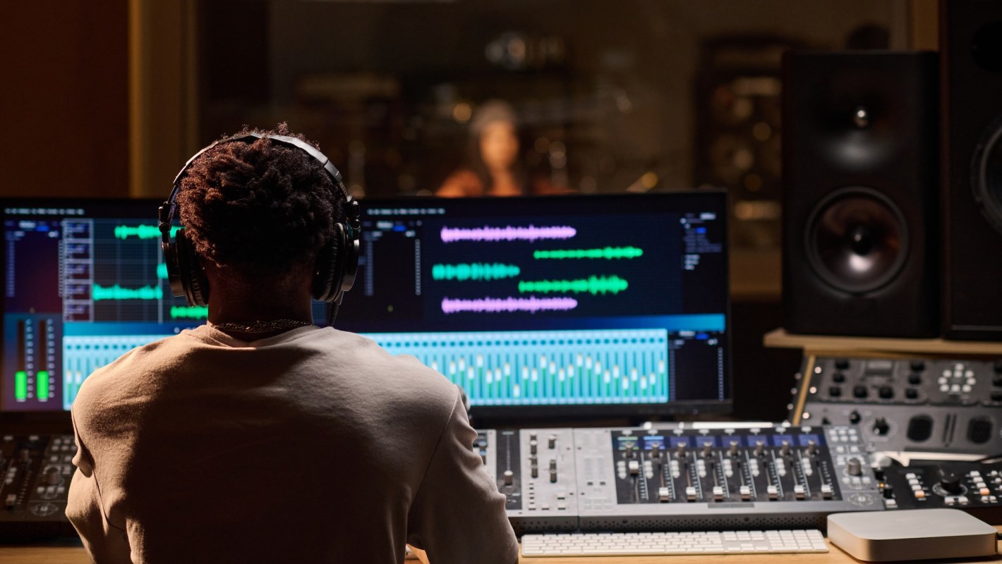 A music producer wearing headphones adjusts settings on a mixing console while monitoring audio waves on a screen.