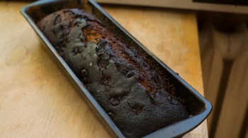 A burnt loaf of bread sits in a black baking pan on a wooden surface.
