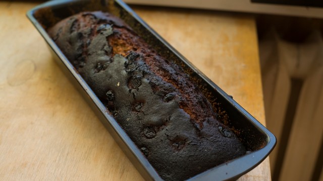A burnt loaf of bread sits in a black baking pan on a wooden surface.