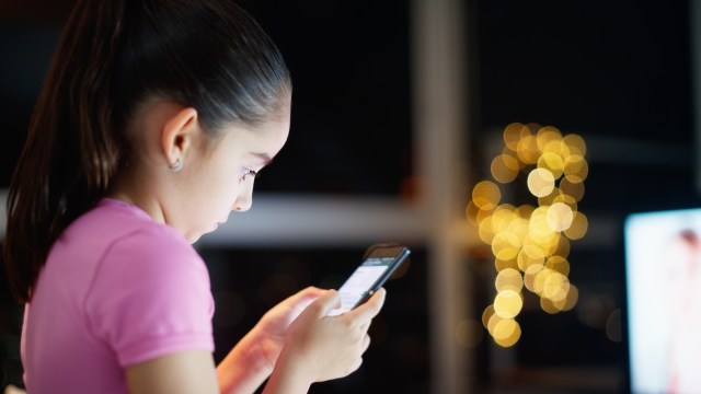 A young girl in a pink shirt focused on her smartphone, with soft bokeh lights in the background.