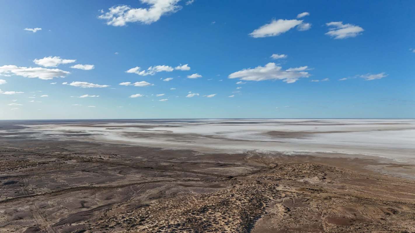 A vast, arid landscape with a salt flat under a bright blue sky dotted with clouds.