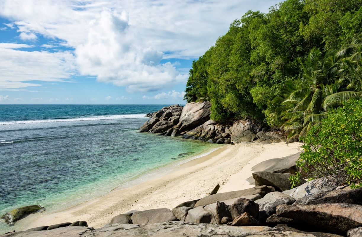 A serene beach with clear turquoise water, rocky formations, and lush greenery under a bright sky.