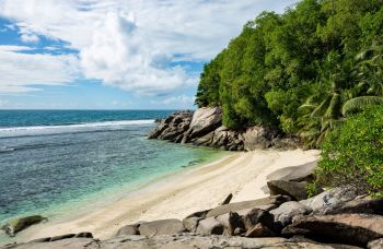 A serene beach with clear turquoise water, rocky formations, and lush greenery under a bright sky.
