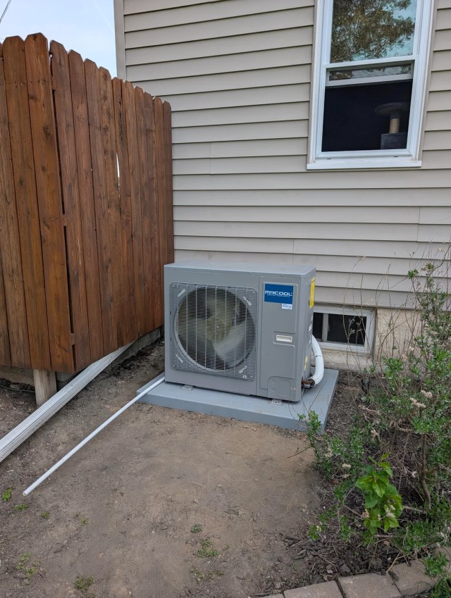 An HVAC is positioned under a window and beside a wooden fence next to a beige house. 