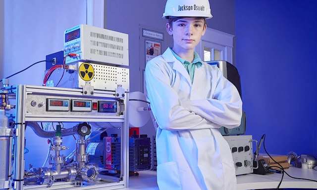 Teen scientist Jackson Oswalt at age 12 in a lab coat and hard hat standing near scientific equipment in a modern lab.