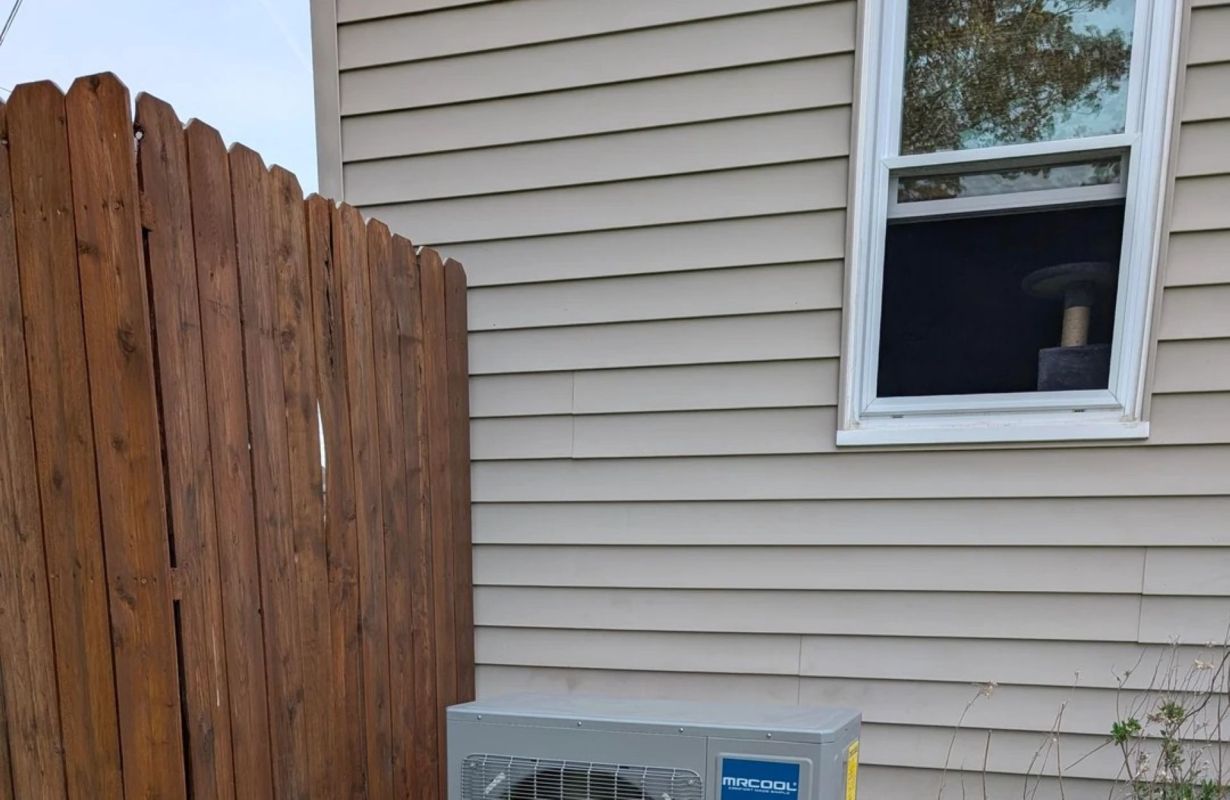 An air conditioning unit is positioned under a window and beside a wooden fence next to a beige house.