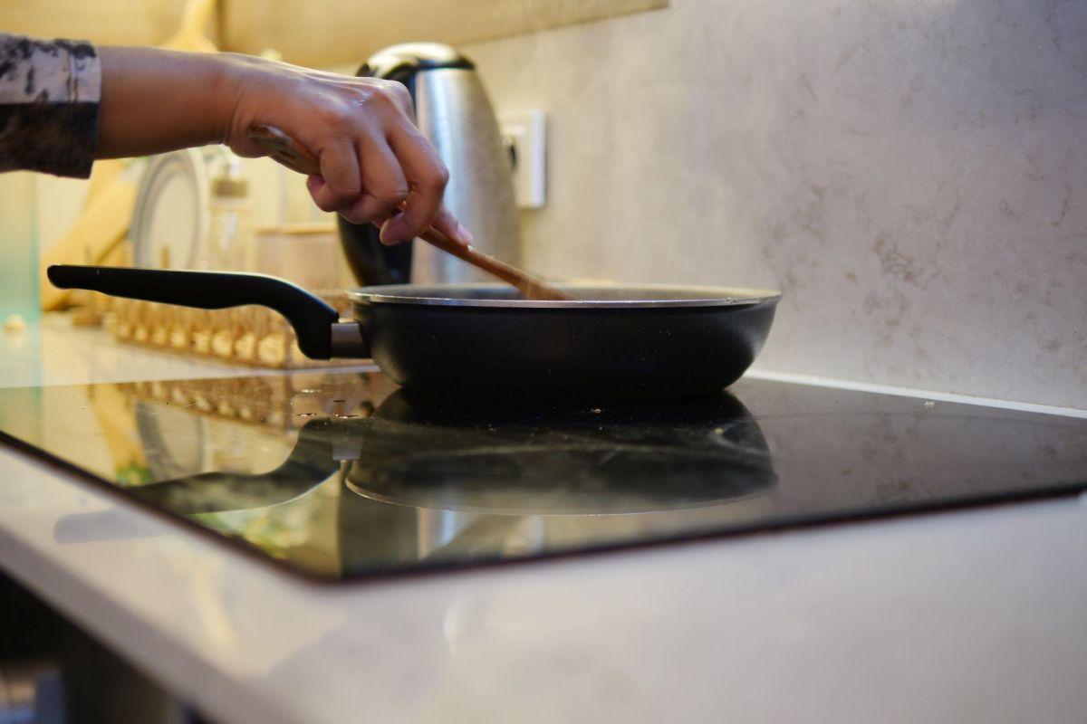 A hand stirring food in a frying pan on a stovetop, with kitchen appliances in the background.