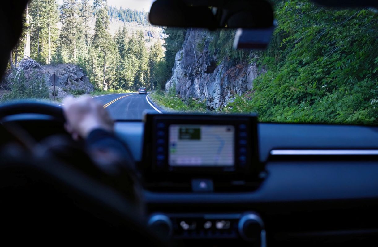 A person's hand on a steering wheel, driving on a winding road surrounded by trees and rocky terrain.