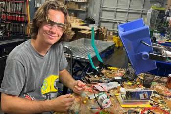 Noah Bild, 19, smiles while working on electronics at a cluttered workshop table filled with tools and materials.