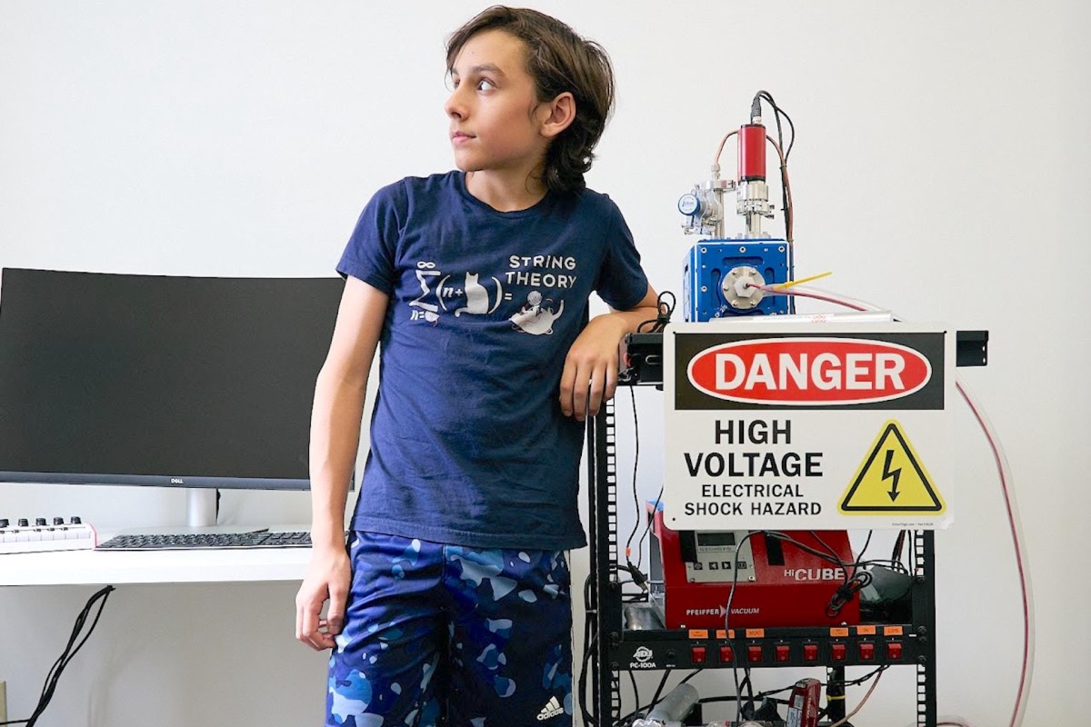 A boy in a "String Theory" t-shirt stands next to a high voltage warning sign and scientific equipment.