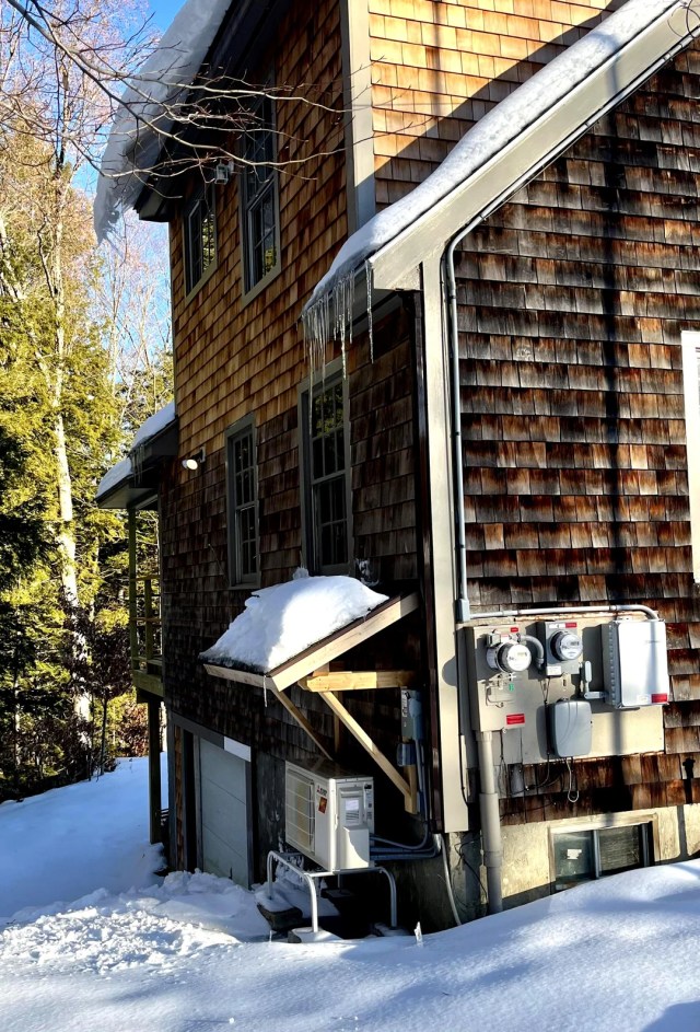 A brown house surrounded by snow, with an additional small overhanging roof constructed to protect a heat pump from the snow.