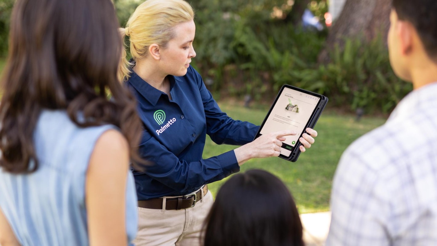 A Palmetto employee in a blue shirt with a tablet talks to two adults and a child.