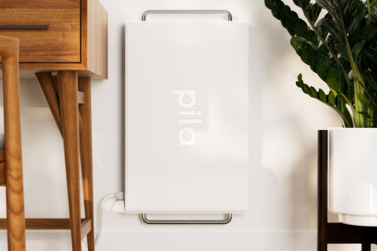A sleek white device labeled "pila" positioned against a wall, next to wooden furniture and a potted plant.