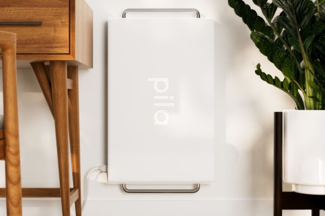 A sleek white device labeled "pila" positioned against a wall, next to wooden furniture and a potted plant.