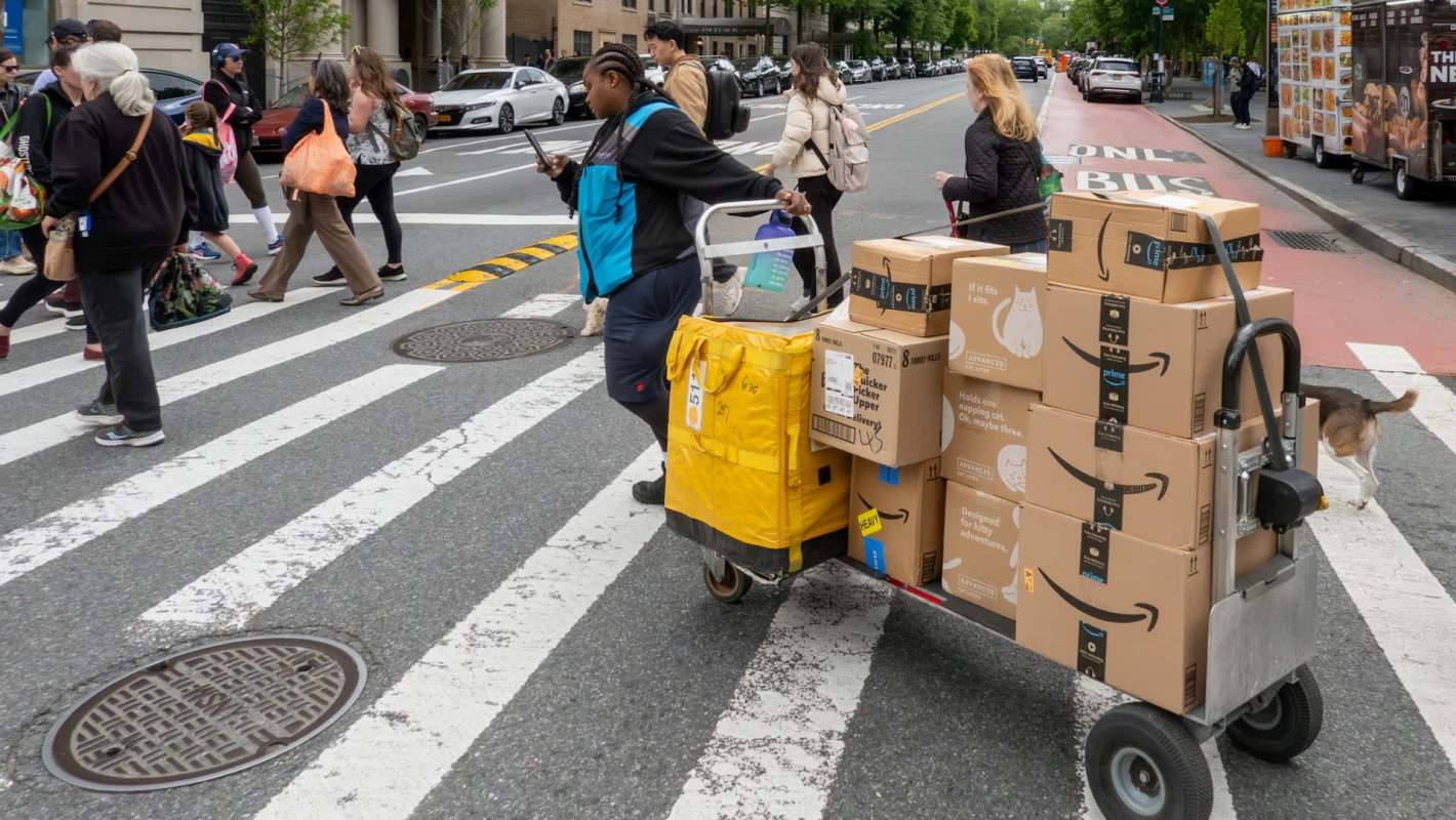 A delivery worker pushes a cart loaded with Amazon packages across a city crosswalk while pedestrians walk by.