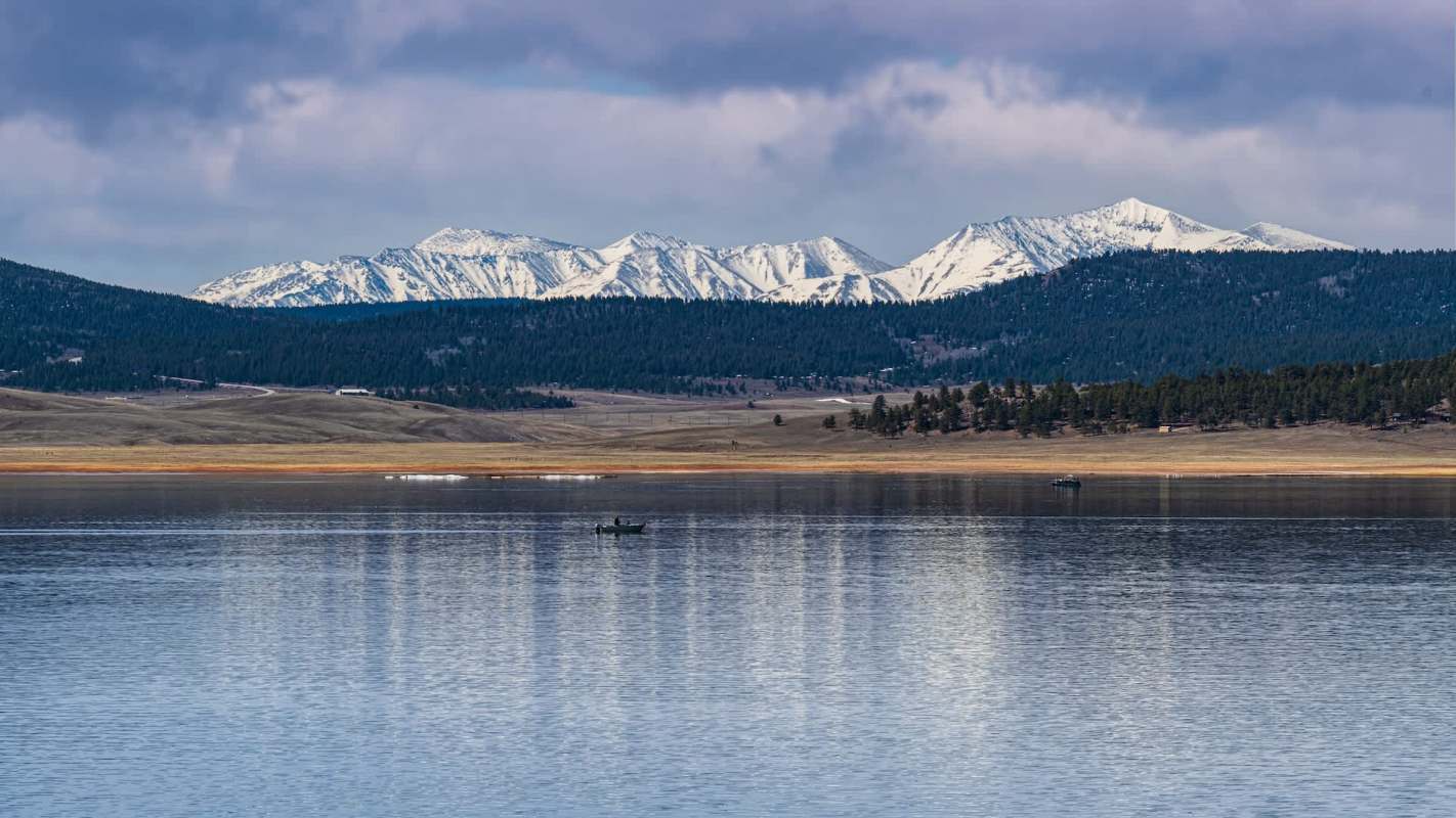 A serene lake with a fisherman in a boat, framed by snow-capped mountains under a cloudy sky.