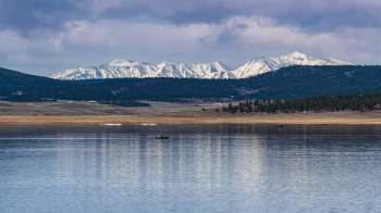 A serene lake with a fisherman in a boat, framed by snow-capped mountains under a cloudy sky.
