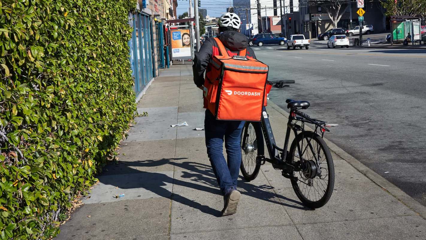 A person in a black jacket walks with a bicycle and an orange DoorDash delivery bag along a city sidewalk.