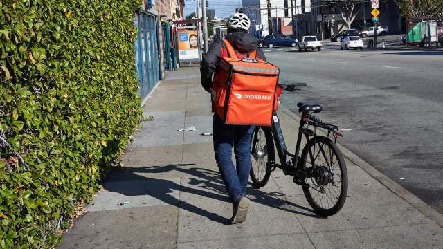 A person walks with a bike and an orange DoorDash delivery bag.