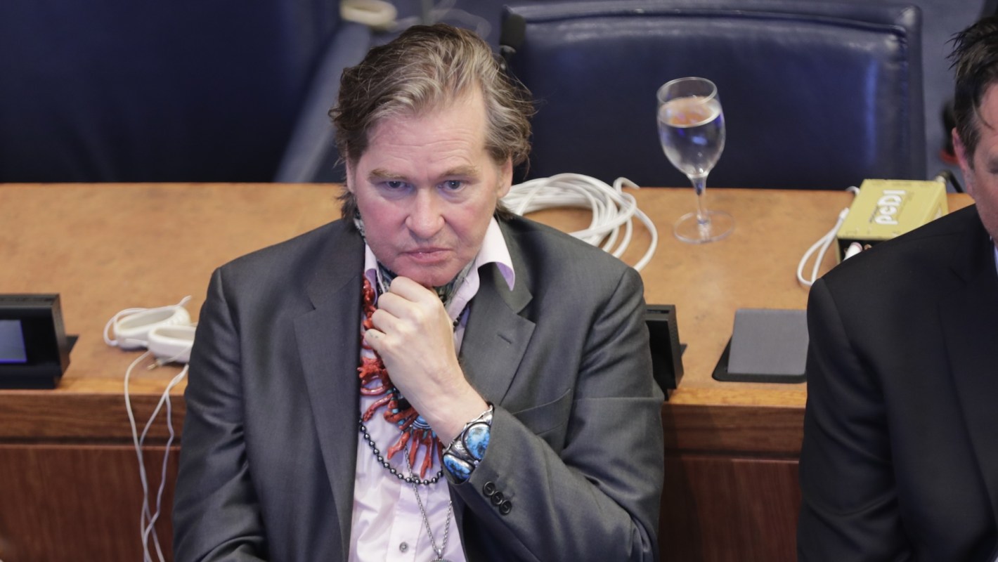 The late actor Val Kilmer, looking contemplative in a suit, sits at a conference table with a glass of water nearby.