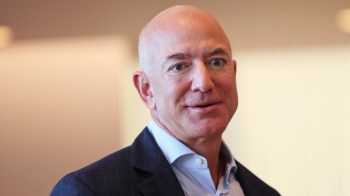 Jeff Bezos, with a shaved head and wearing a navy blazer, speaks with a neutral expression against a blurred background.