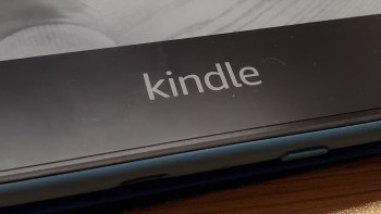 A close-up of a Kindle e-reader logo on a black background with a wooden surface underneath.