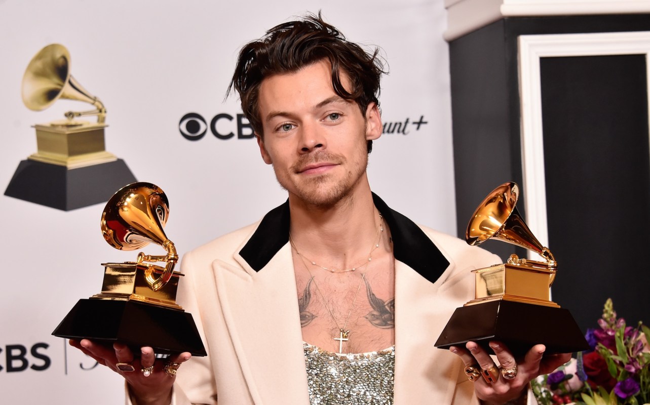 Harry Styles, in a stylish outfit, holds two Grammy Awards, posing confidently in front of a branded backdrop.