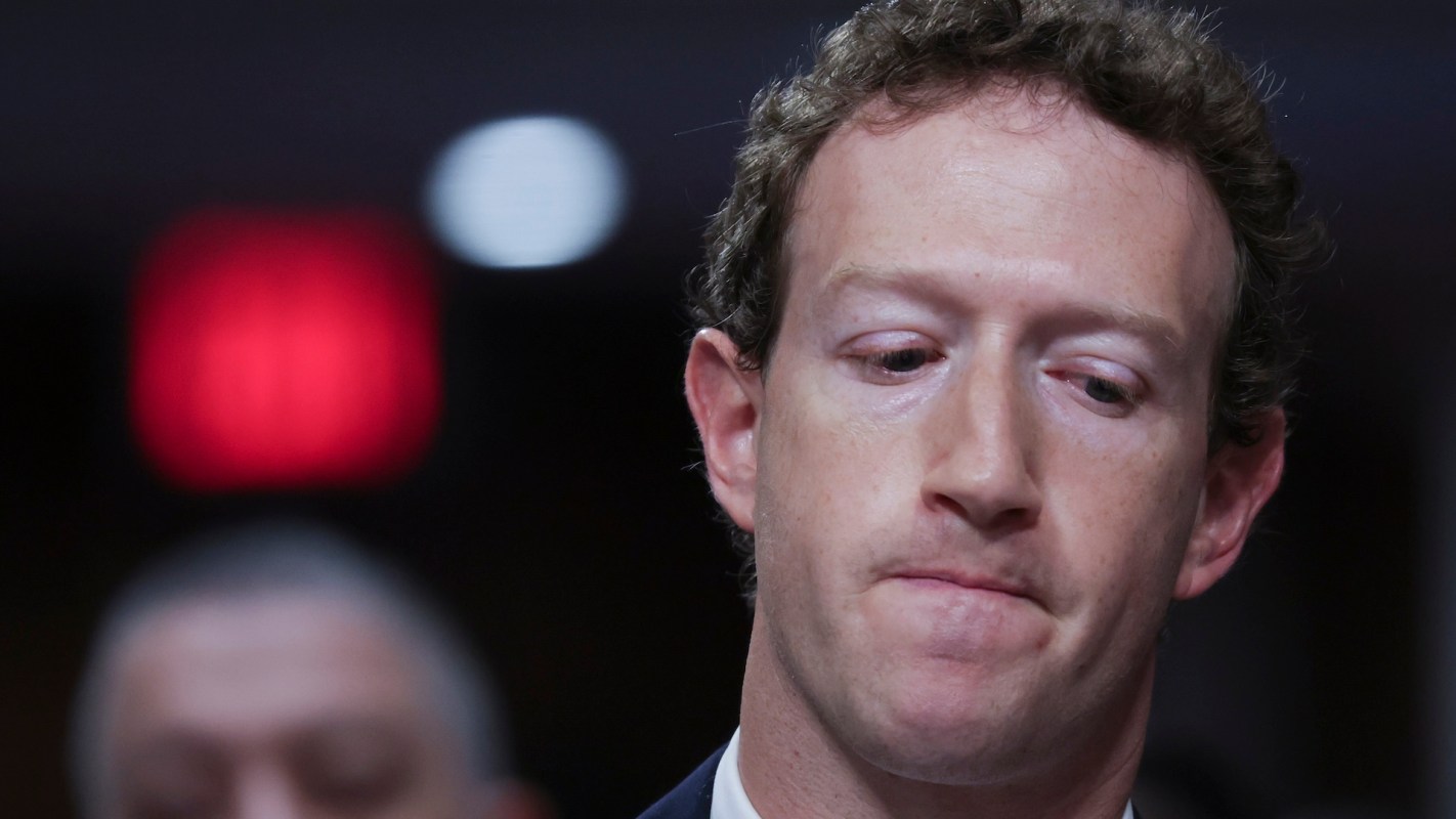 A close-up of Mark Zuckerberg looking down with a serious expression, with blurred figures in the background.