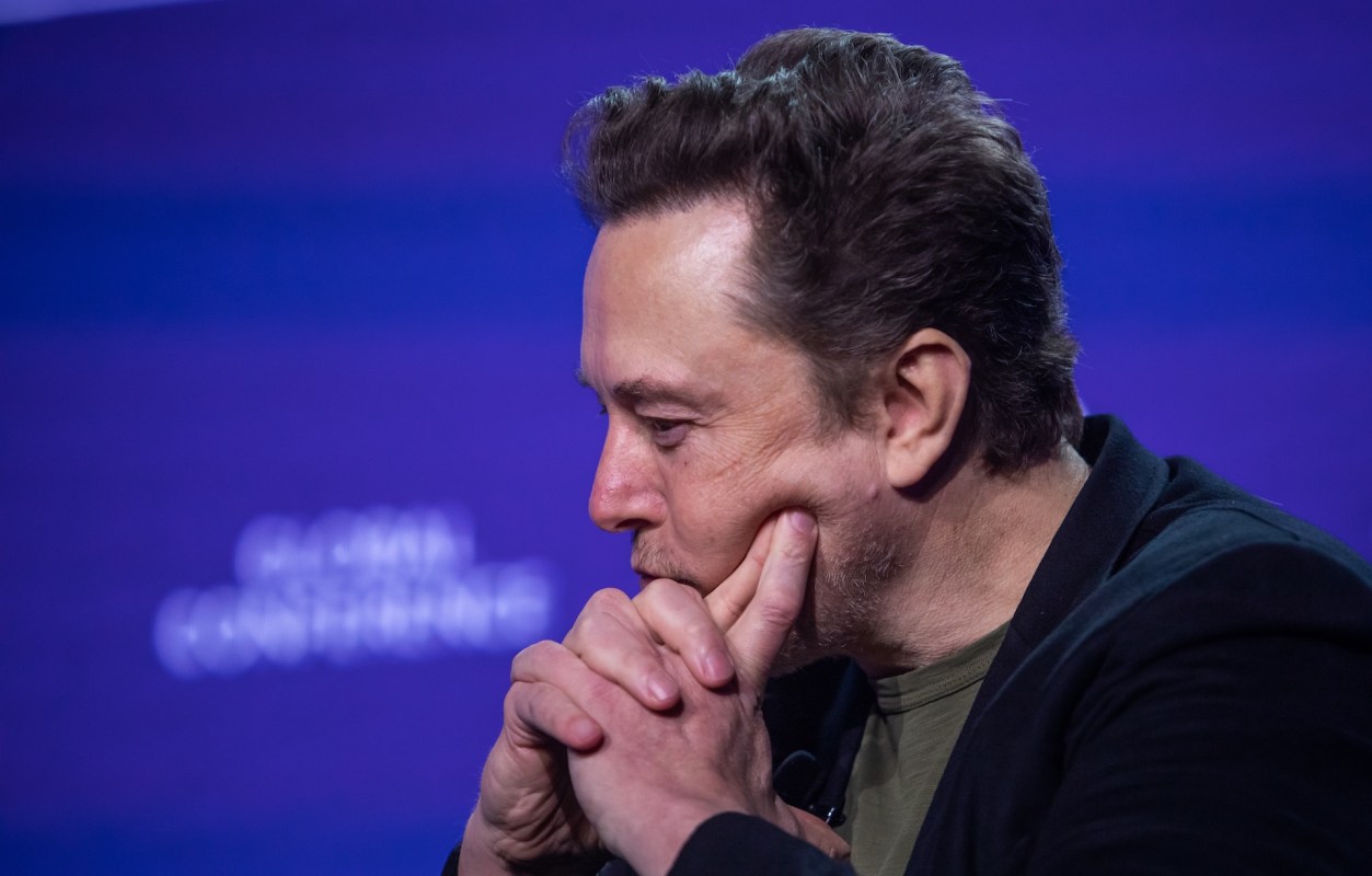 Elon Musk, with short hair and dressed in black, rests his chin on his hands while deep in thought against a purple backdrop.