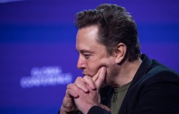 Elon Musk, with short hair and dressed in black, rests his chin on his hands while deep in thought against a purple backdrop.