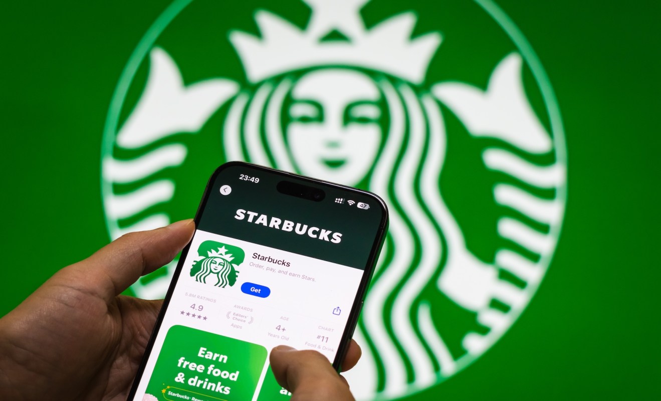 A person holds a smartphone displaying the Starbucks app with a large logo in the background.